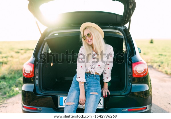 Woman\
sitting in back of car smiling. Getting ready to go. Young laughing\
woman sitting in the open trunk of a car. Summer road trip. Young\
woman sitting in the car trunk with\
suitcases