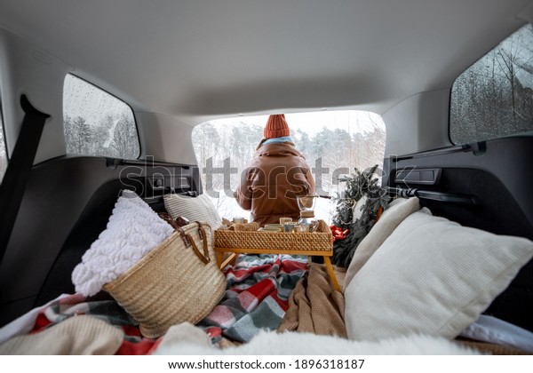Woman sitting back in car trunk near a pine forest,\
traveling by car during winter holidays. View from the backside.\
High quality photo