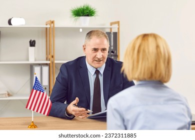 Woman sitting back in the office of the US public services or embassy having consular visa interview with an elderly mature officer with US flag at the desk of official's workplace. - Shutterstock ID 2372311885