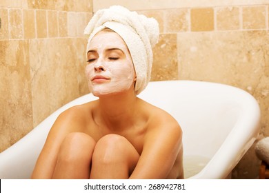 Woman Sitting In Bath With Face Mask, Wearing Towel On Head.