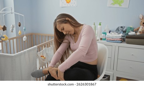 Woman sitting in bedroom holding her leg in pain beside a baby cradle amidst soft toys and gentle light.