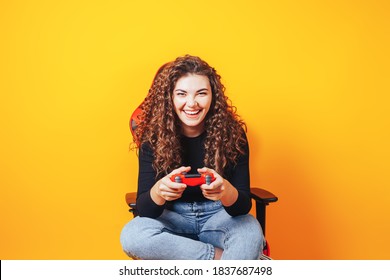 Woman Sitting Behind Gaming Chair In Her Hands Holding Red Gamepad On Yellow Background.