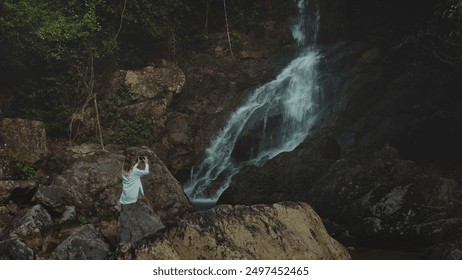 Woman sitting big stone, take picture on mobile phone of waterfall cascade rocky cliff. Female in white shirt relax enjoy beautiful wild nature landscape exotic rainforest. Travel tropical Bali island - Powered by Shutterstock