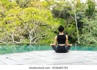 A woman sitting by the edge of a serene infinity pool, surrounded by lush greenery. She is in a meditative pose, enjoying the tranquil atmosphere of nature. The scene conveys peace and relaxation. - Powered by Shutterstock