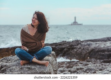 woman sitting by rocky sea beach in wet jeans lighthouse on background. windy weather. summer vacation. carefree concept
