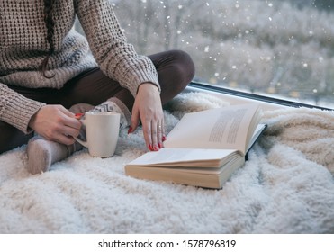 Woman Sitting By The Window Reading Book Drinking Coffee. Winter Snowing Landscape Outside