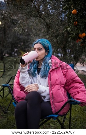 woman sitting at the campsite in cold weather and consuming a beverage from her thermos, outdoor photos of marginal and young woman sitting in green natural environment
