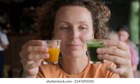 A Woman Sitting In A Catering Establishment And Tasting Two Drinks. Evaluates. One Drink Is Carrot-colored, The Other Is Green. The Woman Has A Satisfied Expression. Drinks To Your Liking.