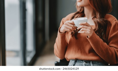 A woman is sitting in a chair with a cup of coffee in her hand. She is wearing an orange sweater and has long brown hair. Concept of relaxation and comfort - Powered by Shutterstock