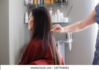 Woman Is Sitting In Chair And Wearing Protective Gown During Treatment Procedures In Beauty Salon. Stylist Is Standing Behind And Straightening Her Long Hair With Comb