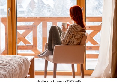 Woman Sitting In A Comfortable Chair In Her Home Drinking Coffee And Looking Through Window At Snow Covered Mountain.