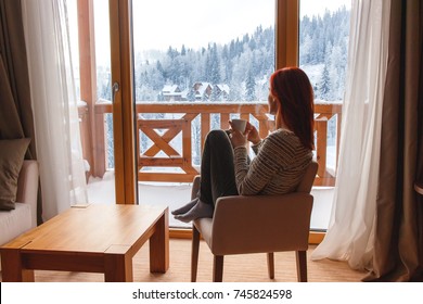 Woman Sitting In A Comfortable Chair In Her Home Drinking Coffee And Looking Through Window At Snow Covered Mountain.