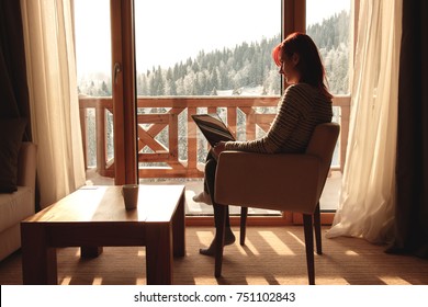 Woman Sitting In A Comfortable Chair In Her Home With Her Laptop And Looking Through Window At Snow Covered Mountain.