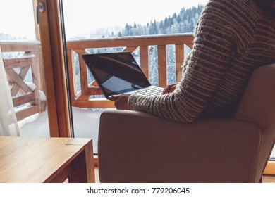 Woman sitting in a comfortable chair with laptop in her home and looking through window at snow covered mountain.