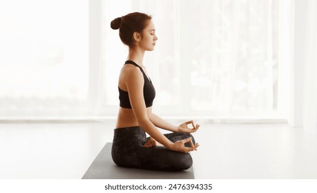 A woman is sitting cross-legged on the floor, practicing yoga. She is focused and holding a pose with her eyes closed, showing concentration and mindfulness, copy space - Powered by Shutterstock