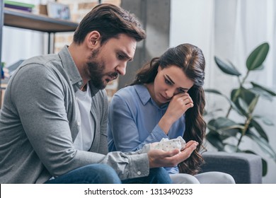 Woman Sitting And Crying On Couch While Man Holding Baby Shoes In Room, Grieving Disorder Concept