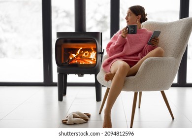 Woman Sitting With Cup On Chair By The Fireplace At Modern House On Nature During Winter Time. Concept Of Winter Mood And Comfort At Home. Girl Wearing Hat And Pink Sweater