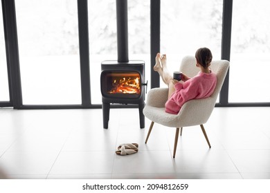 Woman Sitting With Cup On Chair By The Fireplace At Modern House On Nature During Winter Time. Concept Of Winter Mood And Comfort At Home. Girl Wearing Hat And Pink Sweater