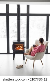 Woman Sitting With Cup On Chair By The Fireplace At Modern House On Nature During Winter Time. Concept Of Winter Mood And Comfort At Home. Girl Wearing Hat And Pink Sweater