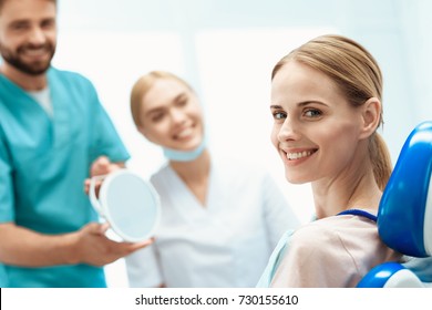 A Woman Is Sitting In A Dental Office In A Dental Chair. In Front Of Her Are Dentists And Are Holding A Mirror. A Woman Looks At The Camera. Happy Patient And Dentist Concept.