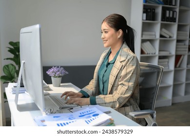 A woman is sitting at a desk with a computer monitor and keyboard in front of her. She is wearing a plaid jacket and a green shirt. The room has a lot of bookshelves and a potted plant
