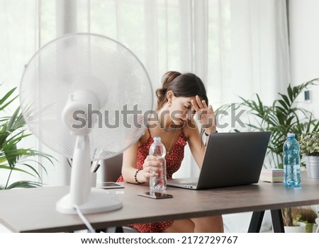 Woman sitting at desk at home during a summer heat wave, she is cooling herself with an electric fan