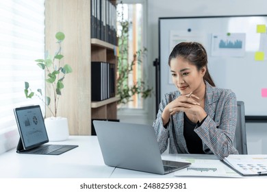 A woman is sitting at a desk with a laptop in front of her. She is wearing a suit and she is focused on her work. The room has a potted plant and a whiteboard on the wall