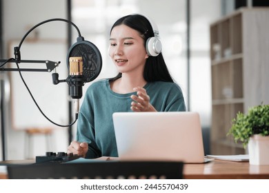 A woman is sitting at a desk with a laptop and a microphone. She is wearing headphones and she is recording a podcast or a voiceover - Powered by Shutterstock