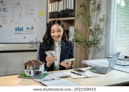 A woman is sitting at a desk with a laptop and a stack of money. She is smiling and she is happy