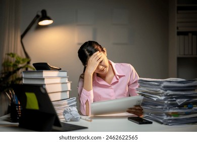 A woman is sitting at a desk with a laptop and a tablet. She is looking at a stack of papers and she is in a state of distress - Powered by Shutterstock