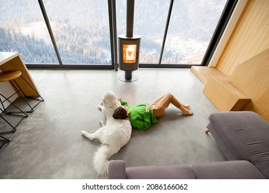 Woman sitting with dog near fireplace and panoramic window at modern living room with stunning view on snowy mountains. Concept of rest in houses or cabins on nature. Wide interior view from above - Powered by Shutterstock