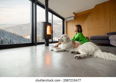 Woman sitting with dog near fireplace and panoramic window at modern living room with stunning view on snowy mountains. Concept of rest in houses or cabins on nature. Idea of escape from everyday life