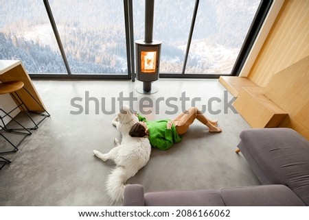 Woman sitting with dog near fireplace and panoramic window at modern living room with stunning view on snowy mountains. Concept of rest in houses or cabins on nature. Wide interior view from above