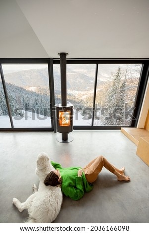 Woman sitting with dog near fireplace and panoramic window at modern living room with stunning view on snowy mountains. Concept of rest in houses or cabins on nature. Wide interior view from above
