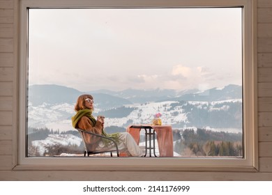 Woman Sitting With A Drink On Terrace Of Tiny House In The Mountains, View Through The Window From The Inside. Concept Of Small Modern Cabins For Rest And Escape To Nature