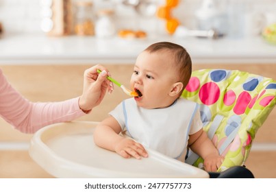 A woman is sitting and feeding a baby with a spoon. The baby is holding onto the spoon and eating from it. The woman is gently guiding the spoon to the baby mouth. - Powered by Shutterstock