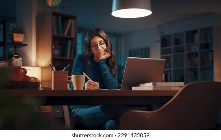 Woman sitting at her desk and connecting with her laptop late at night, she is studying and attending online classes - Powered by Shutterstock