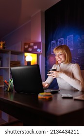 Woman Sitting At Her Desk In Home Office Drinking Coffee And Using Laptop Computer While Working Late At Night