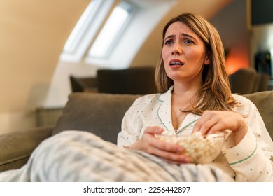A woman is sitting in her pajamas on the couch in the living room she's sad or uneasy, holding popcorn in her lap, she is covered with a blanket and is enjoying her day off and watching movies - Shutterstock ID 2256442897