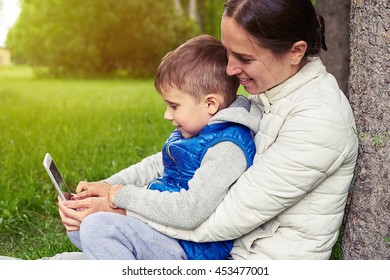 Woman is sitting with her son under the tree in park on a sunny day and watching something on smart phone