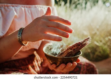 A woman is sitting holding a bowl with a burning smudge stick. The smoke is blowing around her hand. She is in a field of long dry grass.