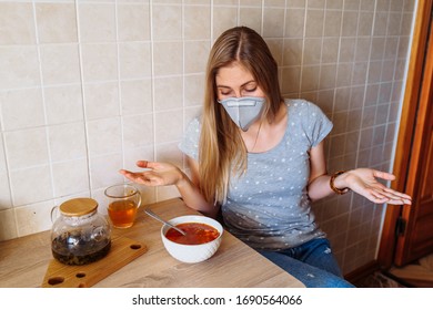 Woman Is Sitting In The Kitchen Near The Table With Food. She Is Isolated At Home During Quarantine Due To The Coronavirus Epidemic, Wearing A Protective Mask.Looking To Plate With Soup And Shrugging