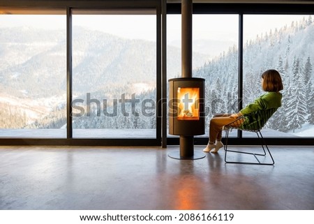 Woman sitting near fireplace at modern living room with great view on snowy mountains. Concept of rest in houses or cabins on nature. Solitude in nature and escape from everyday life