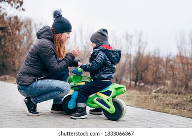 A Woman Is Sitting Near A Little Son Who Rides On A Children's Motorcycle In The Spring. Mom Explains Son Driving Rules On The Street