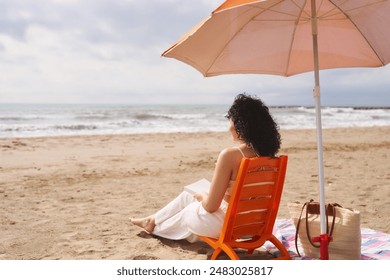 A woman is sitting on a beach chair under an umbrella reading a book. The scene is peaceful and relaxing, with the sound of waves in the background. The woman is enjoying her time alone - Powered by Shutterstock