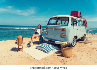 Woman Is Sitting On The Beach For A Picnic Near A Vintage Car, Next To An Old TV Set. Summer Holidays, Road Trip, Vacation, Travel And People Concept - Smiling Young Women Next A Vintage Minivan Car