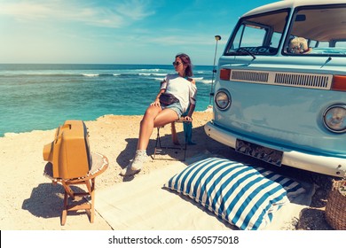 Woman Is Sitting On The Beach For A Picnic Near A Vintage Car, Next To An Old TV Set. Summer Holidays, Road Trip, Vacation, Travel And People Concept - Smiling Young Women Next A Vintage Minivan Car