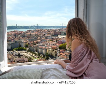 Woman Sitting On The Bed In The Hotel Room And Looking At View From Window At Lisbon City Streets