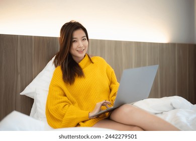 A woman is sitting on a bed with a laptop in front of her. She is wearing a yellow sweater and is using the laptop. Concept of relaxation and comfort - Powered by Shutterstock