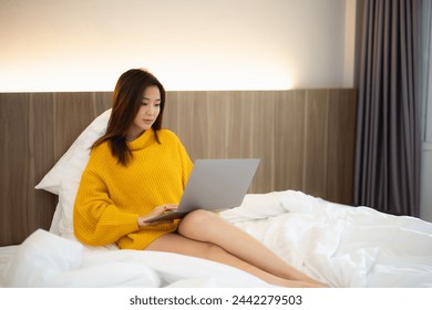 A woman is sitting on a bed with a laptop in front of her. She is wearing a yellow sweater and is using the laptop. Concept of relaxation and comfort - Powered by Shutterstock